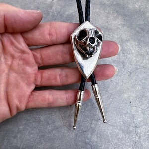 May include: A silver bolo tie with a skull pendant. The skull is detailed and has a pointed shape. The bolo tie is on a black cord.