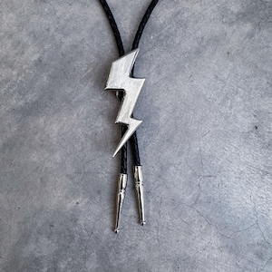 Op de afbeelding: Een bolo-das met een zilverkleurige bliksemschicht-hanger in het midden van een zwart gevlochten koord. De uiteinden van het koord zijn afgewerkt met gepolijste zilverkleurige metalen punten, wat een strakke uitstraling geeft aan dit accessoire.