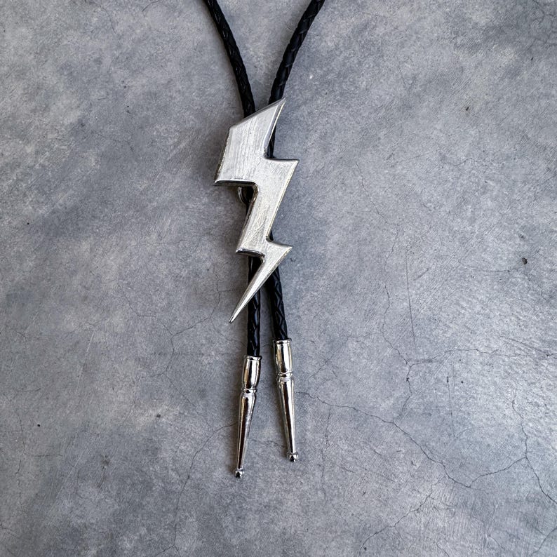 Op de afbeelding: Een bolo-das met een zilverkleurige bliksemschicht-hanger in het midden van een zwart gevlochten koord. De uiteinden van het koord zijn afgewerkt met gepolijste zilverkleurige metalen punten, wat een strakke uitstraling geeft aan dit accessoire.
