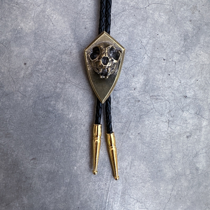 May include: A black bolo tie with a gold-toned pendant featuring a detailed animal skull. The pendant is shaped like a shield.