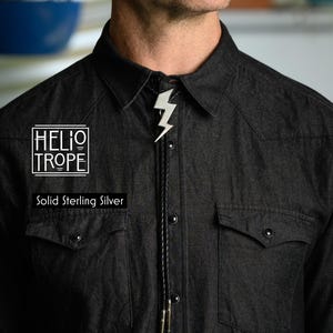 Op de afbeelding: Een bolo-das met een bliksemschicht-hanger van massief sterling zilver aan een zwart gevlochten koord met metalen uiteinden. De tekst HELIOTROPE en Solid Sterling Silver is zichtbaar aan de linkerkant van het donkergrijze overhemd.