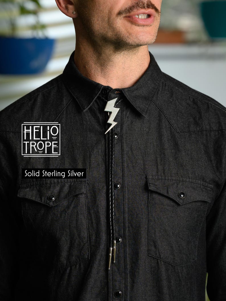 Op de afbeelding: Een bolo-das met een bliksemschicht-hanger van massief sterling zilver aan een zwart gevlochten koord met metalen uiteinden. De tekst HELIOTROPE en Solid Sterling Silver is zichtbaar aan de linkerkant van het donkergrijze overhemd.