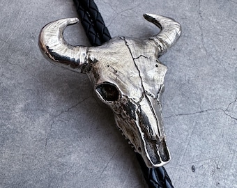 Op de afbeelding: Een bolo-das met een zilverkleurige metalen stierenschedelhanger met gedetailleerde hoorn- en botstructuren. De hanger is bevestigd aan een zwart gevlochten leren koord dat verticaal door de achterkant van de schedel loopt.
