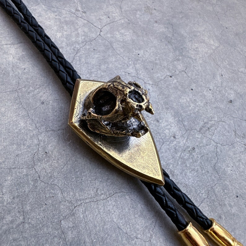 May include: A gold bolo tie with a skull-shaped pendant. The pendant is detailed and has a textured surface. The tie is made of black leather cord.