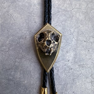 May include: A gold bolo tie with a detailed skull pendant. The skull is set in a shield-shaped frame and has a textured surface.