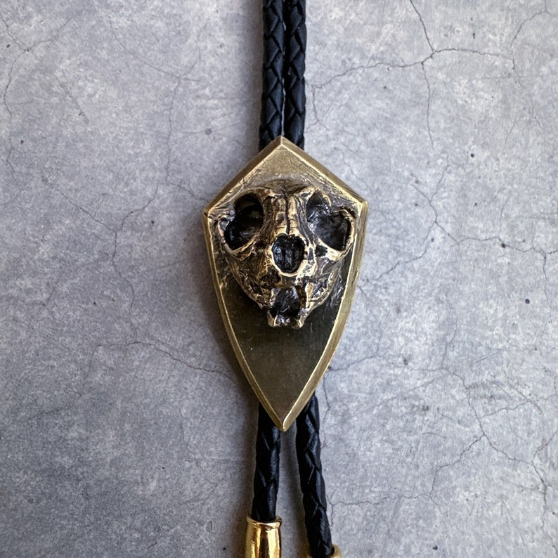 May include: A gold bolo tie with a detailed skull pendant. The skull is set in a shield-shaped frame and has a textured surface.