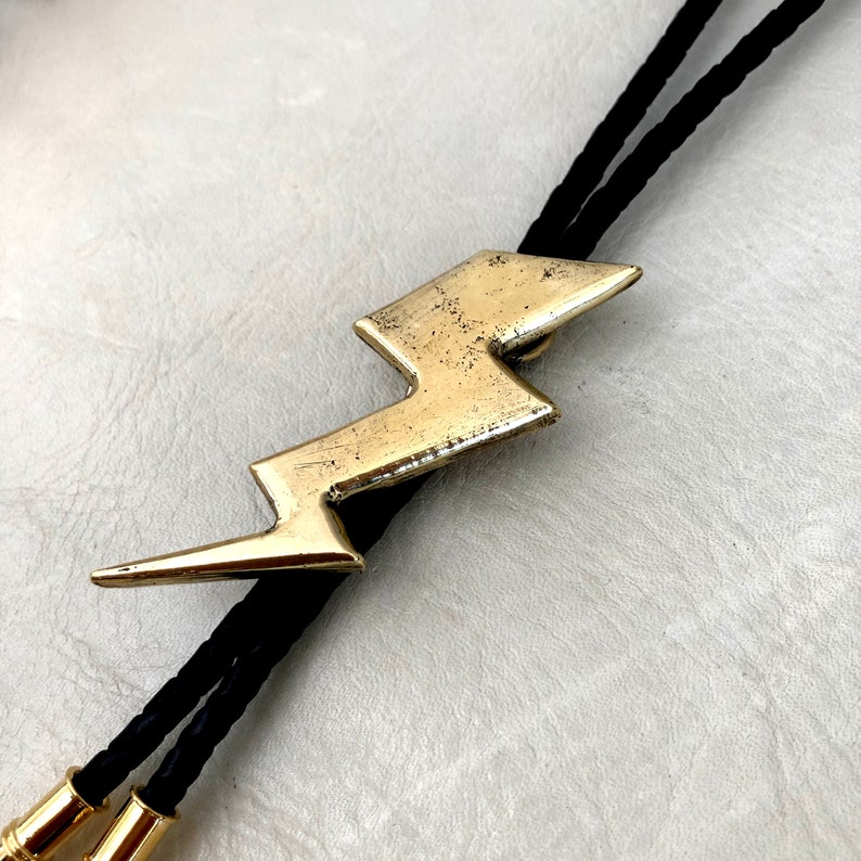 Op de afbeelding: Een bolo-das met een goudkleurige metalen bliksemschicht-hanger met een verweerde, versleten afwerking. De hanger is bevestigd aan een zwart gevlochten koord dat eindigt in twee gepolijste goudkleurige metalen uiteinden, een opvallend nekaccessoire.