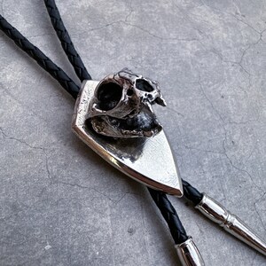 May include: A silver bolo tie with a skull pendant. The skull is detailed and has a textured surface. The bolo tie is made of black leather cord.