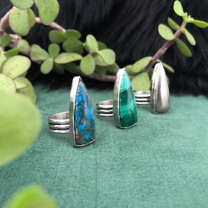 May include: Three silver rings with different colored gemstones. The rings are on a green surface. The gemstones are turquoise, green, and silver.