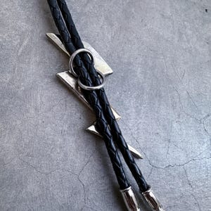 Op de afbeelding: Een bolo-das met een zilverkleurige bliksemschicht-hanger. Het koord is gemaakt van zwart gevlochten leer en wordt op zijn plaats gehouden door een ronde zilverkleurige schuif. De twee uiteinden van het leren koord zijn afgewerkt met gepolijste zilverkleurige metalen punten.