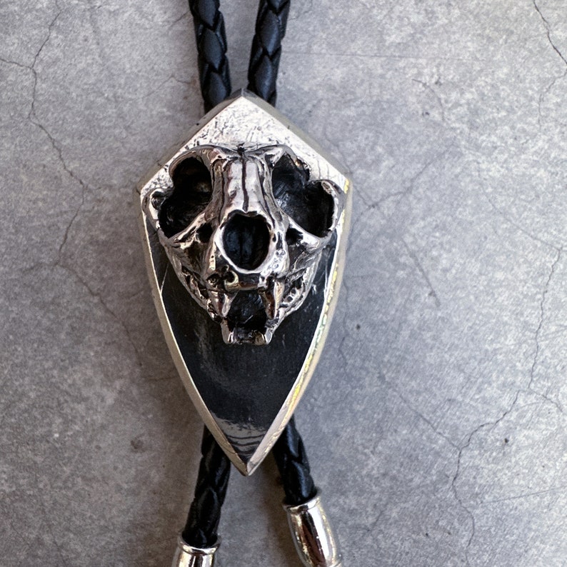 May include: A silver bolo tie with a detailed skull pendant. The skull is set in a shield-shaped frame and has a black background.