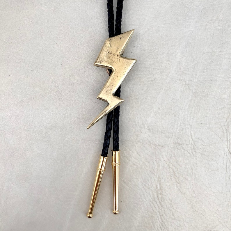 Op de afbeelding: Een bolo-das met een goudkleurige bliksemschicht-hanger met een verweerde textuur. De hanger is bevestigd aan een zwart gevlochten koord dat eindigt in twee bijpassende goudkleurige metalen uiteinden aan de onderkant van het koord.
