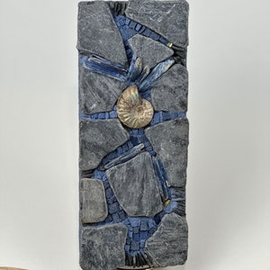 May include: A rectangular slate tile mosaic with a blue and gray color scheme. The mosaic features a fossilized ammonite shell in the center, surrounded by smaller pieces of slate and blue glass.