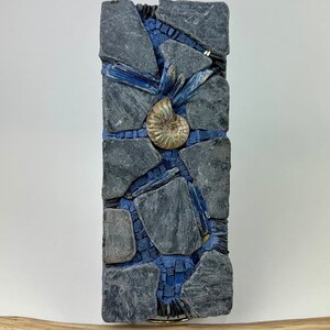 May include: A mosaic wall art piece featuring a blue and gray color scheme. The mosaic is made of small pieces of slate and glass. A fossilized ammonite is featured in the center of the piece.