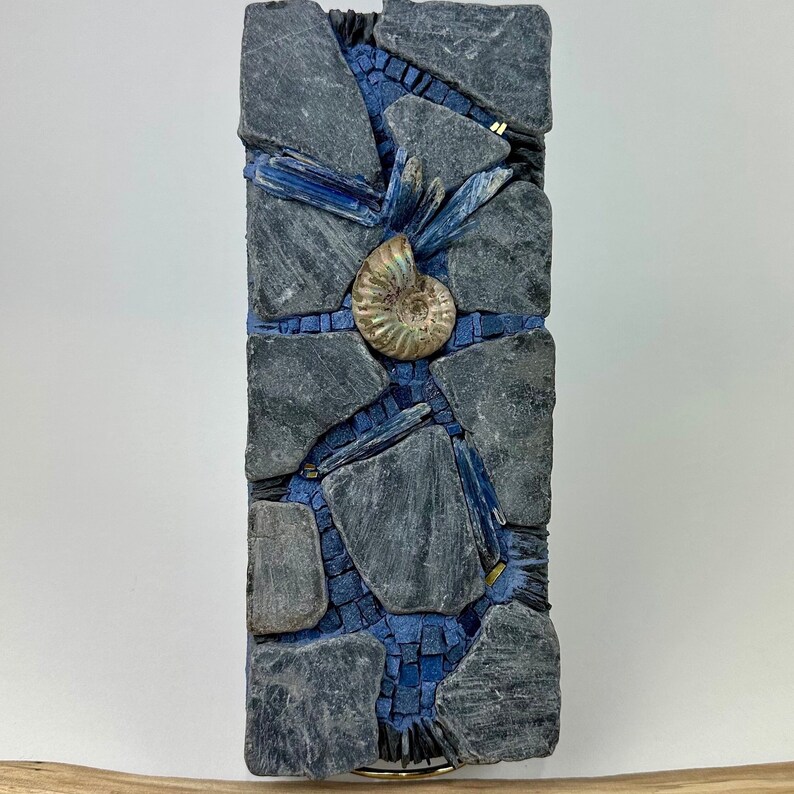May include: A mosaic wall art piece featuring a blue and gray color scheme. The mosaic is made of small pieces of slate and glass. A fossilized ammonite is featured in the center of the piece.