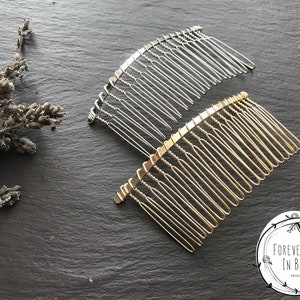 May include: Two hair combs, one silver and one gold, on a dark grey surface. The combs are made of metal and have a curved shape. The combs are both approximately 10 cm long.