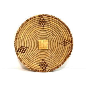 May include: A round woven basket with a brown and tan pattern. The basket is made of natural materials and has a circular shape.