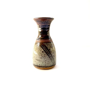 May include: A small, hand-thrown ceramic vase with a speckled glaze in shades of brown, gray, and white. The vase has a narrow neck and a rounded body.