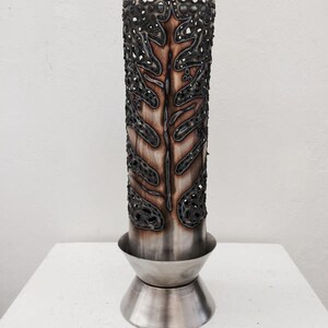 Puede incluir: Un jarrón de metal plateado con un diseño decorativo de hoja recortada. El jarrón tiene una base redonda.