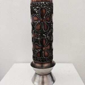 Puede incluir: Un jarrón de cobre con un patrón floral decorativo cortado en el metal. El jarrón tiene una base plateada.