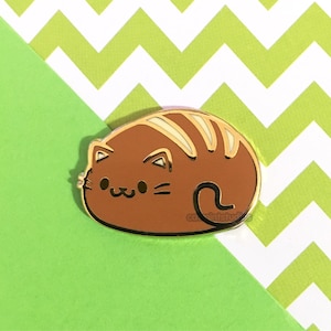 May include: A brown cat enamel pin with gold trim, featuring a cute, cartoonish design. The cat has cream-colored stripes and a smiling face. The pin is set against a green and white chevron background.