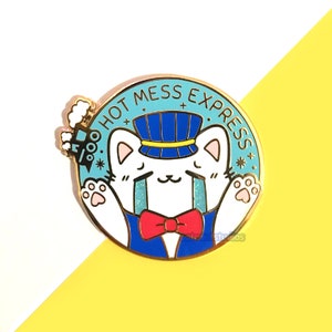May include: A gold-colored enamel pin featuring a white cat wearing a blue conductor's hat and bow tie. The pin has the text "HOT MESS EXPRESS" in a blue circle, with a small train on the left.