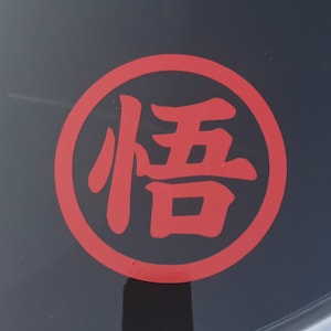 May include: A red circular sticker with Japanese characters on a car window. The sticker features a bold, red design with a black background. The Japanese character is in the center of the circle.
