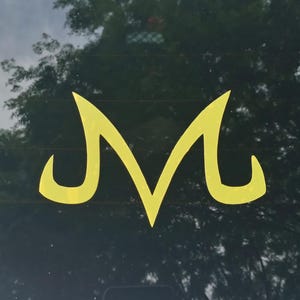 May include: A yellow vinyl decal featuring the Majin symbol, characterized by its distinct M-shape with sharp, pointed top edges and curved, hook-like bottom terminals. The sticker is applied to the center of a dark, reflective glass surface.