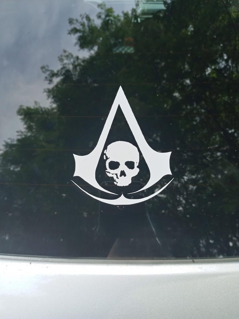 May include: A white vinyl decal applied to a glass surface, featuring a stylized triangular emblem with a central skull graphic. The design consists of sharp, pointed edges and a hollowed-out skull silhouette centered within the geometric shape.