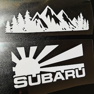 May include: Two white vinyl decals on a black backing. The top decal features a silhouette of a mountain range with pine trees. The bottom decal displays a stylized sunburst graphic positioned above the word SUBARU in bold white capital letters.