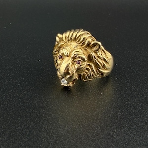 May include: A gold-colored lion head ring with a detailed mane and a diamond accent in its mouth. The lion's eyes are small red gems. The ring is set against a dark background.