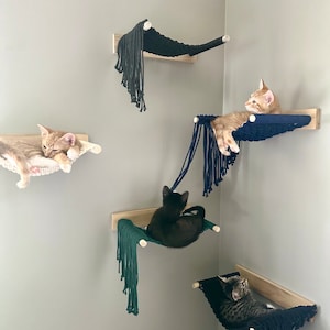 May include: Five cat beds made of woven rope are mounted on the wall. The beds are in a corner of the room and are all different colors. There are four cats in the beds, one is black, one is orange, and two are tabby.