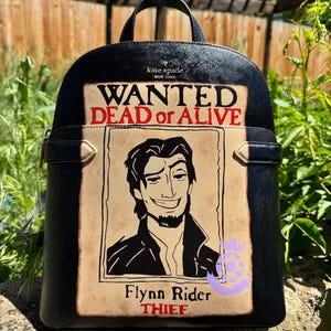 以下が含まれることがあります： 「Wanted Dead or Alive」のポスターデザインが特徴の黒いバックパック。ポスターには、「Flynn Rider, Thief」という名前の笑顔の男性が描かれています。バックパックには上部のハンドルとKate Spade New Yorkのロゴが付いています。