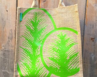 May include: A brown jute tote bag with green palm tree designs. The bag has two handles and is hanging on a wooden surface.