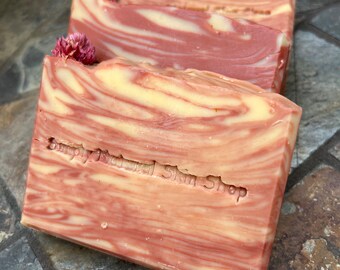 Puede incluir: Tres barras de jabón hechas a mano con un diseño en espiral en tonos rosa y crema. Las barras están rematadas con pequeñas flores rosas y tienen el texto "Simply Natural Skin Shop" grabado en la parte delantera.