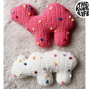 May include: Two crocheted animal plushies shaped like frosted animal crackers. The pink plush is a camel and the white plush is a bear. Both plushies have colorful sprinkles on them.