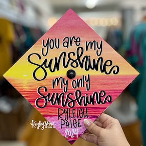 May include: A graduation cap with a sunset design, featuring the text "you are my sunshines, my only sunshines." The cap includes the name "Ryleigh Paige" and the year "2024."