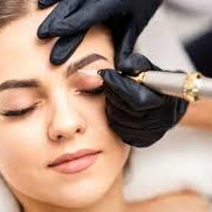 Puede incluir: Primer plano del rostro de una persona durante un procedimiento de microblading de cejas. Un técnico con guantes negros utiliza una herramienta manual para aplicar pigmento en la zona de las cejas. Los ojos de la persona están cerrados.