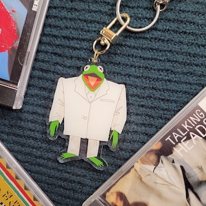 May include: A green frog wearing a white suit with a gold chain keychain attached. The keychain is hanging from a silver chain.