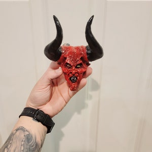 Puede incluir: Una cabeza de diablo roja con cuernos negros, el diablo tiene una expresión amenazante con dientes afilados y ojos amarillos. La cabeza de diablo está hecha de un material duro y está pintada en un estilo realista.