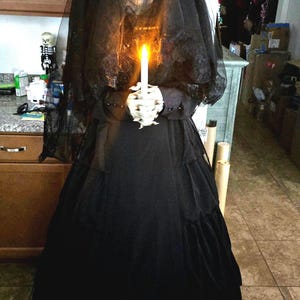 May include: A life-sized figure dressed in a long black gown and a matching black veil. The figure has a pale, skeletal face with dark eye sockets and a wide, toothy grin. It holds a single, upright, glowing orange candle in its pale hands.