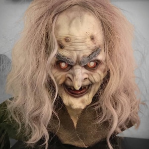 Puede incluir: Un busto realista y detallado de una bruja con cabello largo y blanco. La bruja tiene una cara arrugada con una nariz grande y torcida y ojos rojos penetrantes. El busto está pintado en un estilo realista y es una gran adición a cualquier colección de Halloween o fantasía.