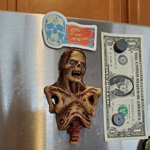 May include: A zombie-themed magnet with a detailed, decaying torso design. The magnet is affixed to a metal surface, alongside a dollar bill and a magnet with the text "Little Corpse Magnets" and a skull graphic.