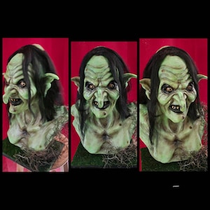 May include: A green-skinned, female goblin bust with long black hair and a pointed nose. The goblin has a mischievous expression with a wide grin and sharp teeth. The bust is made of a realistic material and is painted in a detailed manner.