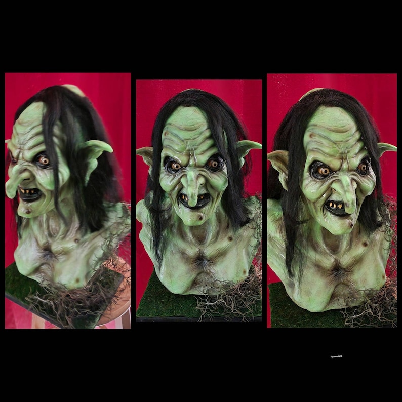 May include: A green-skinned, female goblin bust with long black hair and a pointed nose. The goblin has a mischievous expression with a wide grin and sharp teeth. The bust is made of a realistic material and is painted in a detailed manner.