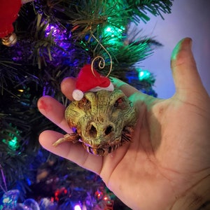 May include: A green and gold Christmas ornament shaped like a dragon's head wearing a red Santa hat.