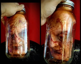 Könnte beinhalten: Ein menschlicher Kopf, der in einem durchsichtigen Glasgefäß mit einem silbernen Deckel aufbewahrt wird. Der Kopf ist mit Blut bedeckt und hat ein realistisches Aussehen.
