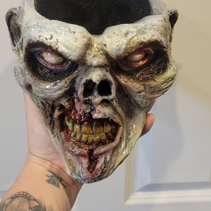 Könnte beinhalten: Ein Keramik-Pflanztopf in Form eines Zombiekopfes mit einem rissigen, blutigen Mund und sichtbaren Zähnen. Der Topf ist in Weiß-, Grün- und Rottönen bemalt.