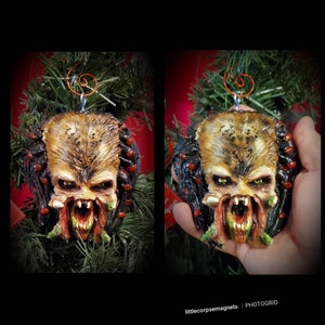 Pode incluir: Um enfeite de Natal com o tema Predator, com um rosto detalhado e ameaçador. O enfeite apresenta uma cabeça marrom e bege com mandíbulas abertas, dentes afiados e detalhes em vermelho e preto. É suspenso por um fio cor de cobre e colocado sobre um fundo verde.