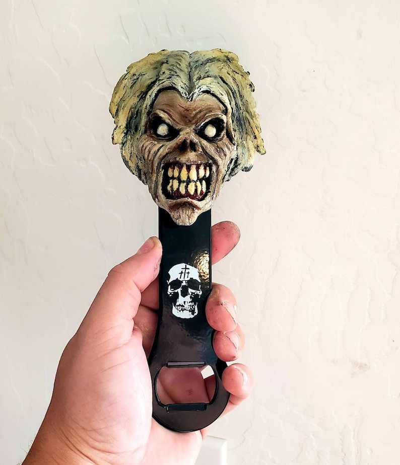 Może przedstawiać: Czarny metalowy otwieracz do butelek z rzeźbioną głową Eddiego, maskotki zespołu heavy metalowego Iron Maiden. Głowa jest pomalowana w odcieniach zieleni, ż&oacute;łci i brązu. Otwieracz do butelek zawiera r&oacute;wnież czarną i białą czaszkę z napisem "Kill" nad nią.